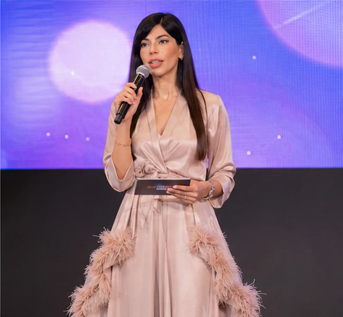 Rima Iskandarani hosting an evening gala in a blush satin feathered gown, holding a microphone before a purple stage