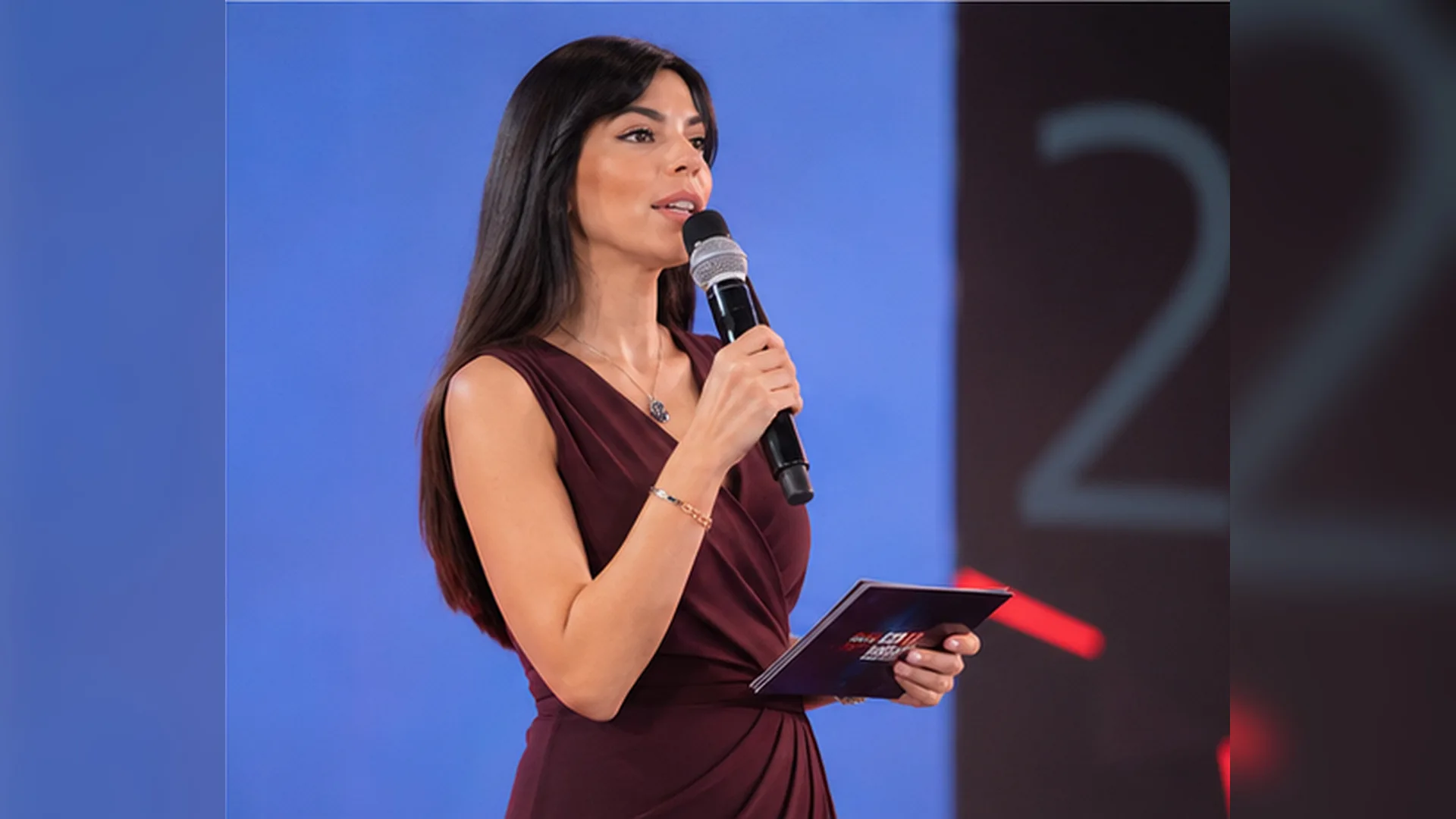 Rima Iskandarani hosting an awards ceremony in a burgundy gown, speaking into a microphone while holding cue cards