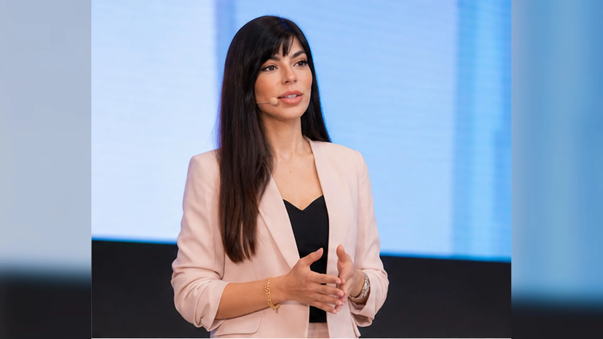 Rima Iskandarani speaking at a conference in a blush blazer with a headset microphone, presenting to the audience