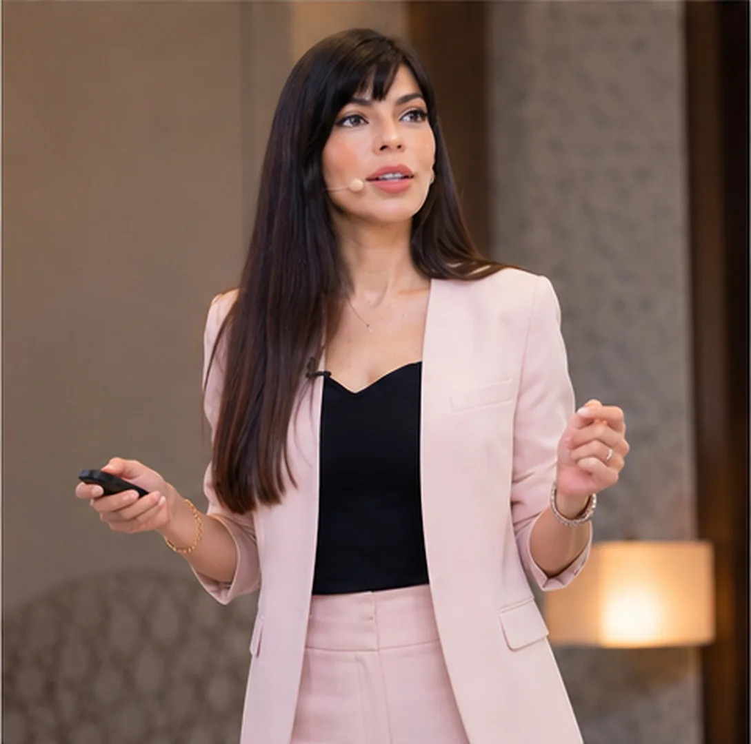 Rima Iskandarani presenting in a blush suit with a headset microphone in a warmly lit venue