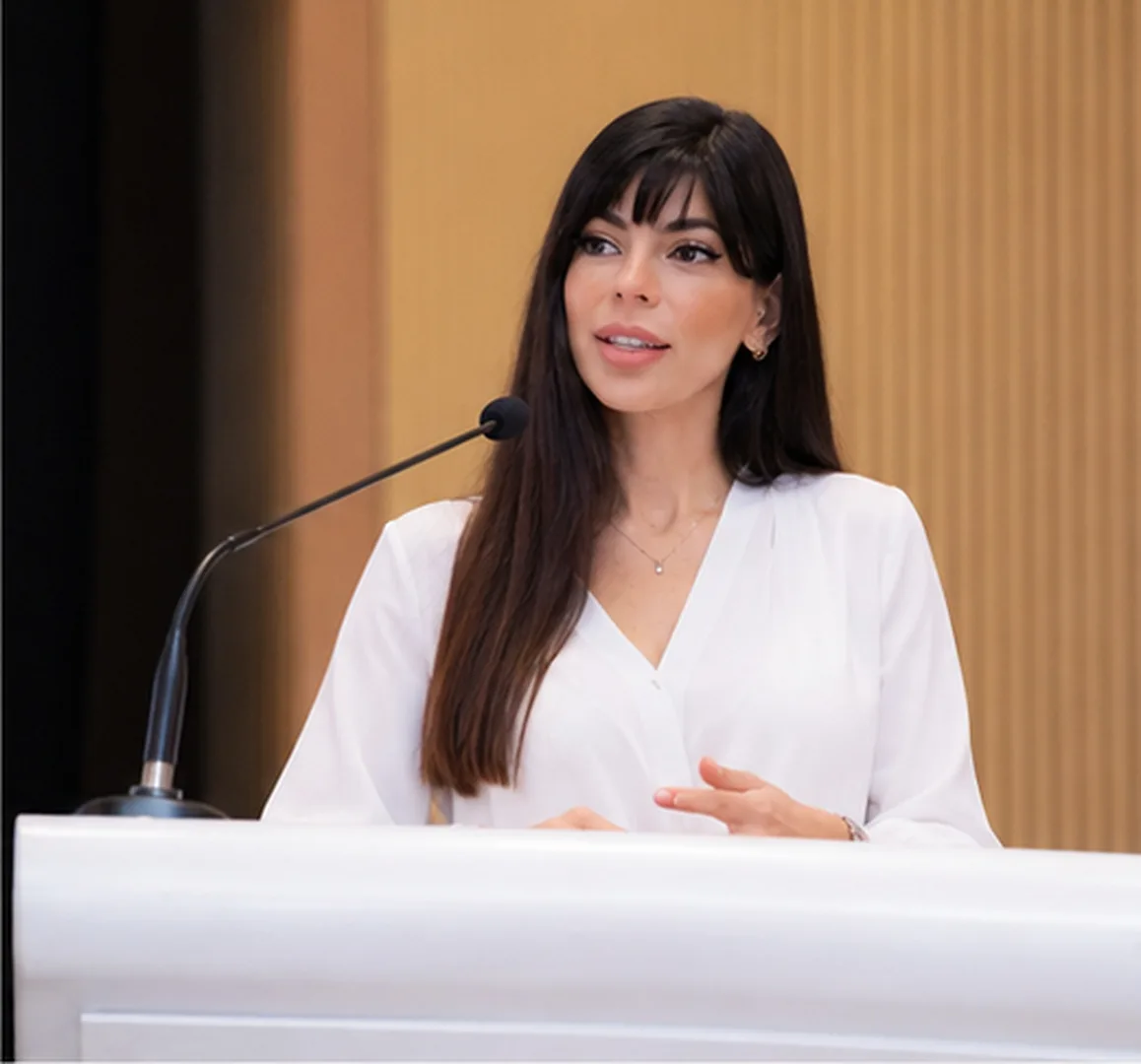Rima Iskandarani delivering an address from a podium in a white blouse against a warm backdrop