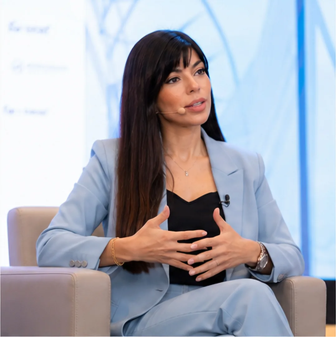 Rima Iskandarani leading a seated panel discussion in a light blue pantsuit, gesturing as she speaks