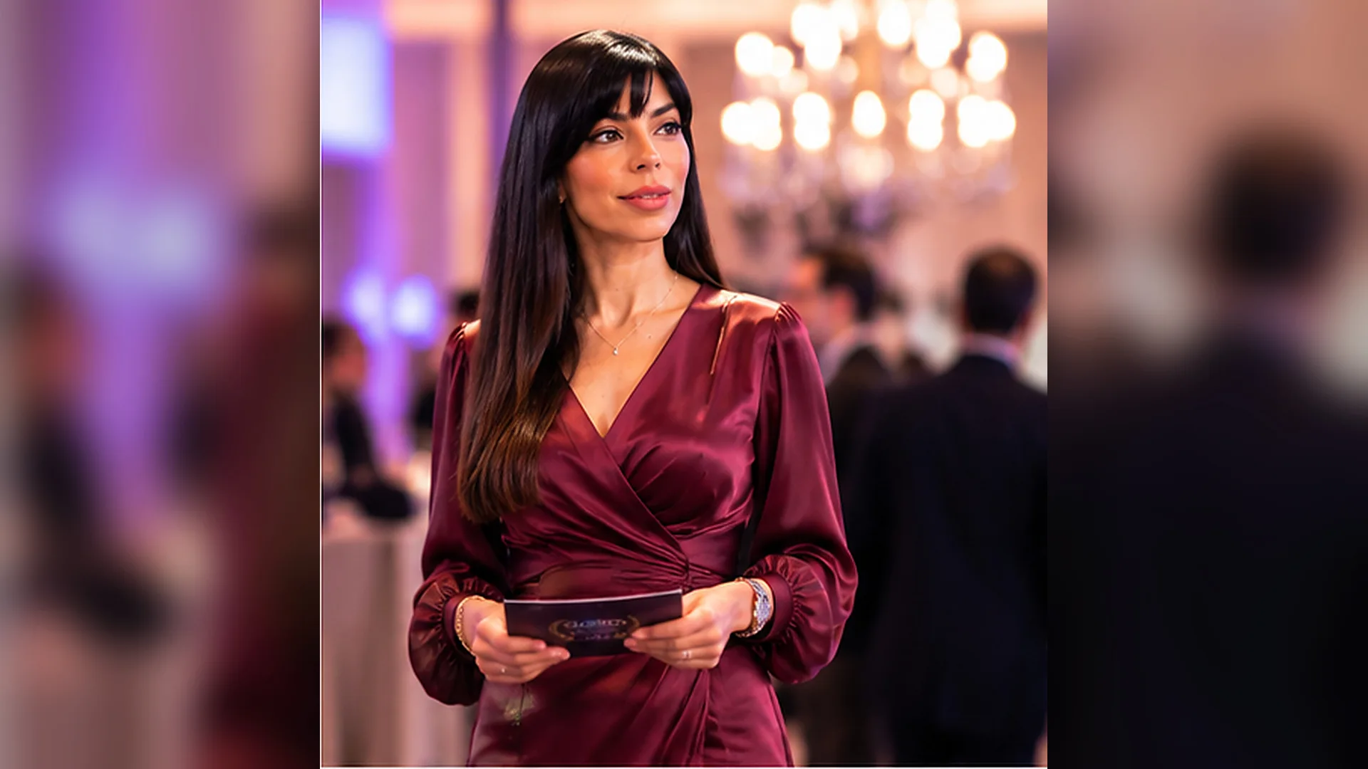 Rima Iskandarani hosting a gala reception in a burgundy satin dress, holding a programme card in a chandelier-lit ballroom
