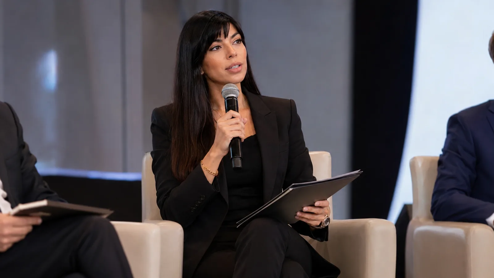 Rima Iskandarani in a finance forum moderation setting, seated with a handheld microphone and notes